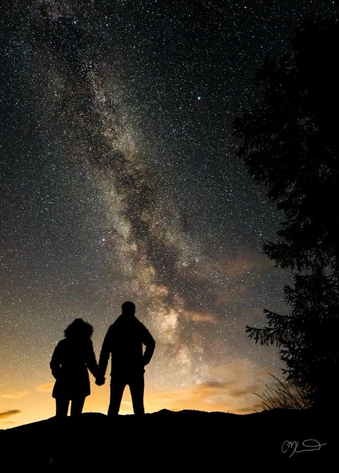 What a breathtaking shot! Two silhouettes stand, looking outward at star dense skies as dusk descends.