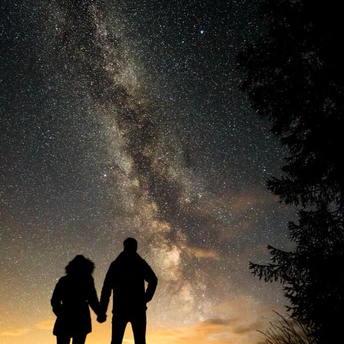 What a breathtaking shot! Two silhouettes stand, looking outward at star dense skies as dusk descends.