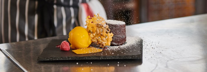 Study Food studies and Hospitality at Perth College UHI. This shows a delicious chocolate souffle with a honeycomb crisp, raspberry and a yellow coloured fruit sorbet served on a slate and being sprinkled with white icing sugar.